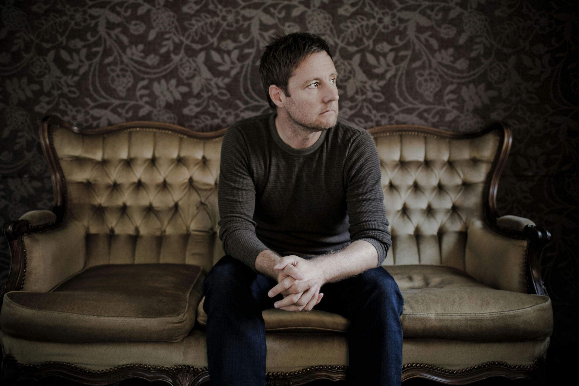 Man sitting on an elegant vintage sofa in a room with intricate wallpaper, looking contemplative.
