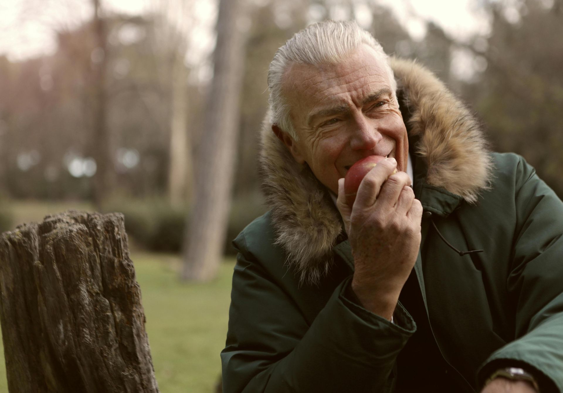 Smiling senior man wearing a green parka while eating an apple outdoors.