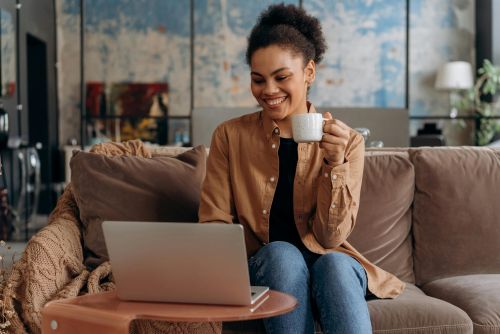 A relaxed woman smiling while working at home with a laptop and coffee.