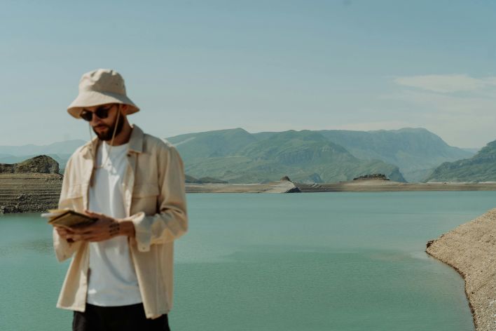 A man wearing a bucket hat and sunglasses looks at a book by a peaceful reservoir.