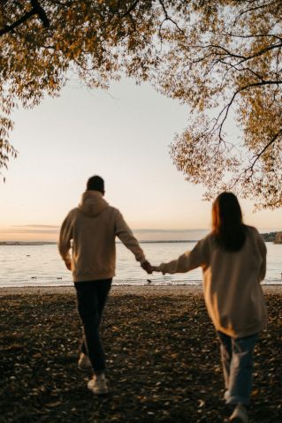 A romantic couple holding hands while walking towards the sunset by the beach, surrounded by autumn scenery.